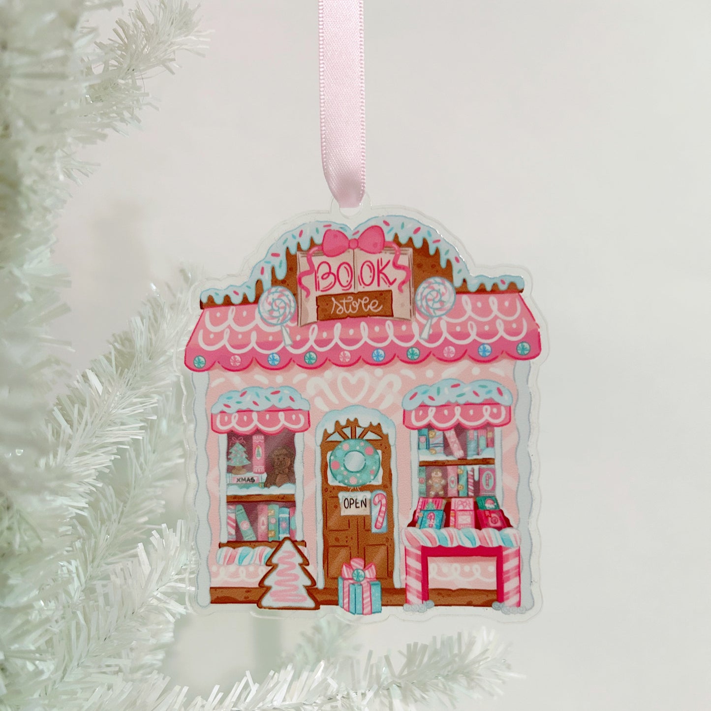 Decorative ornament shaped like a bookshop with a pink roof, hanging on a white Christmas tree.