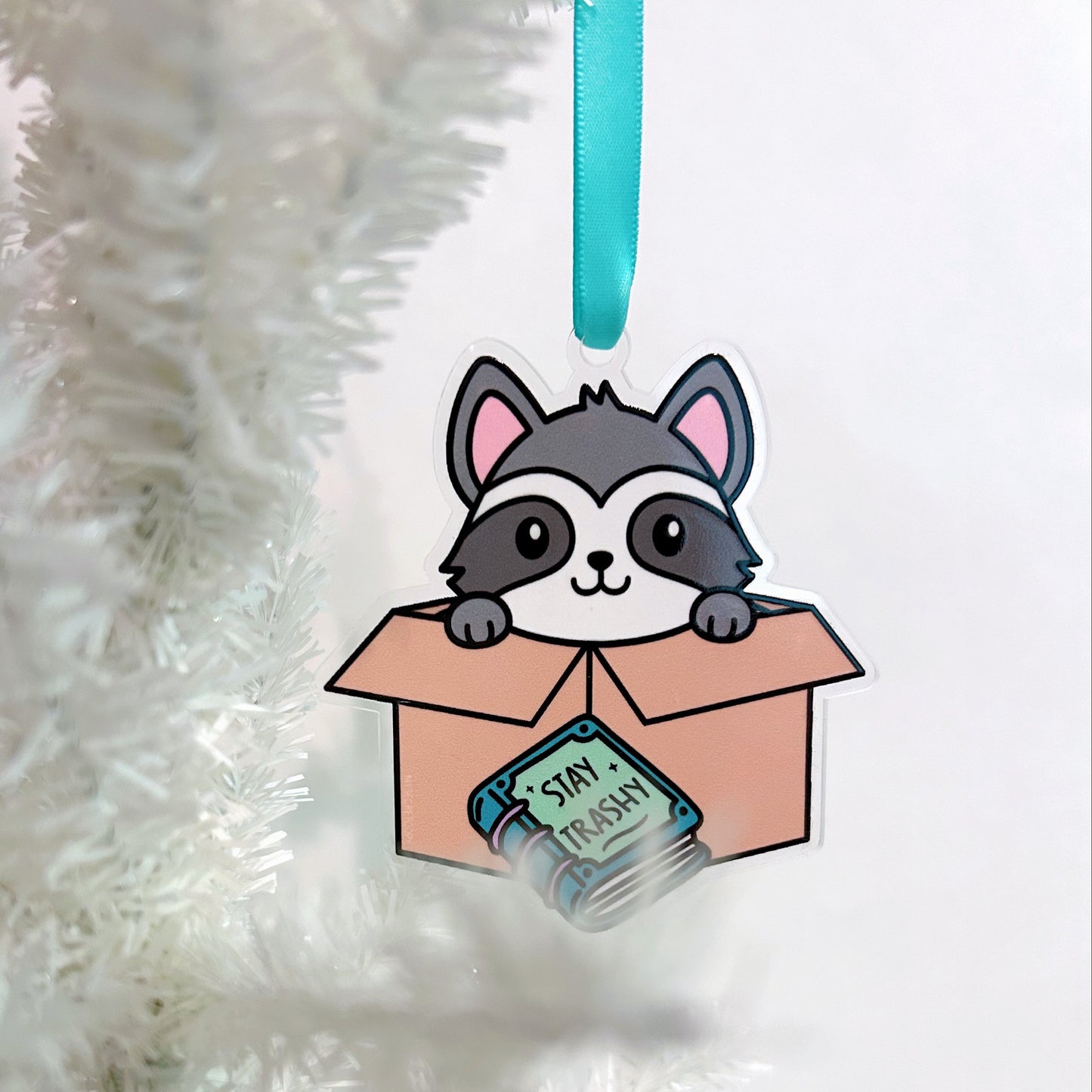 Decorative ornament with a raccoon in a box and book that says "stay trashy" on a white Christmas tree.