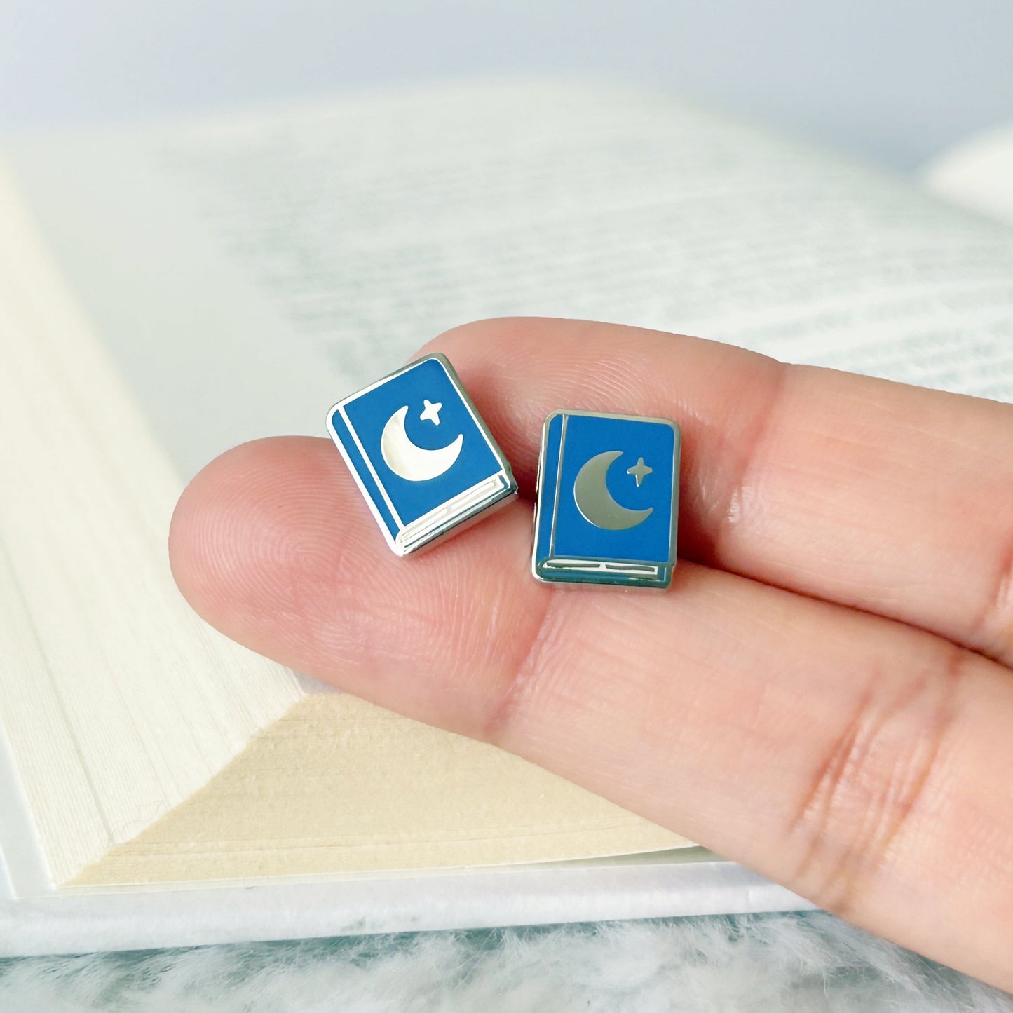 Blue square earrings with a moon and star design held between fingers on a light background.