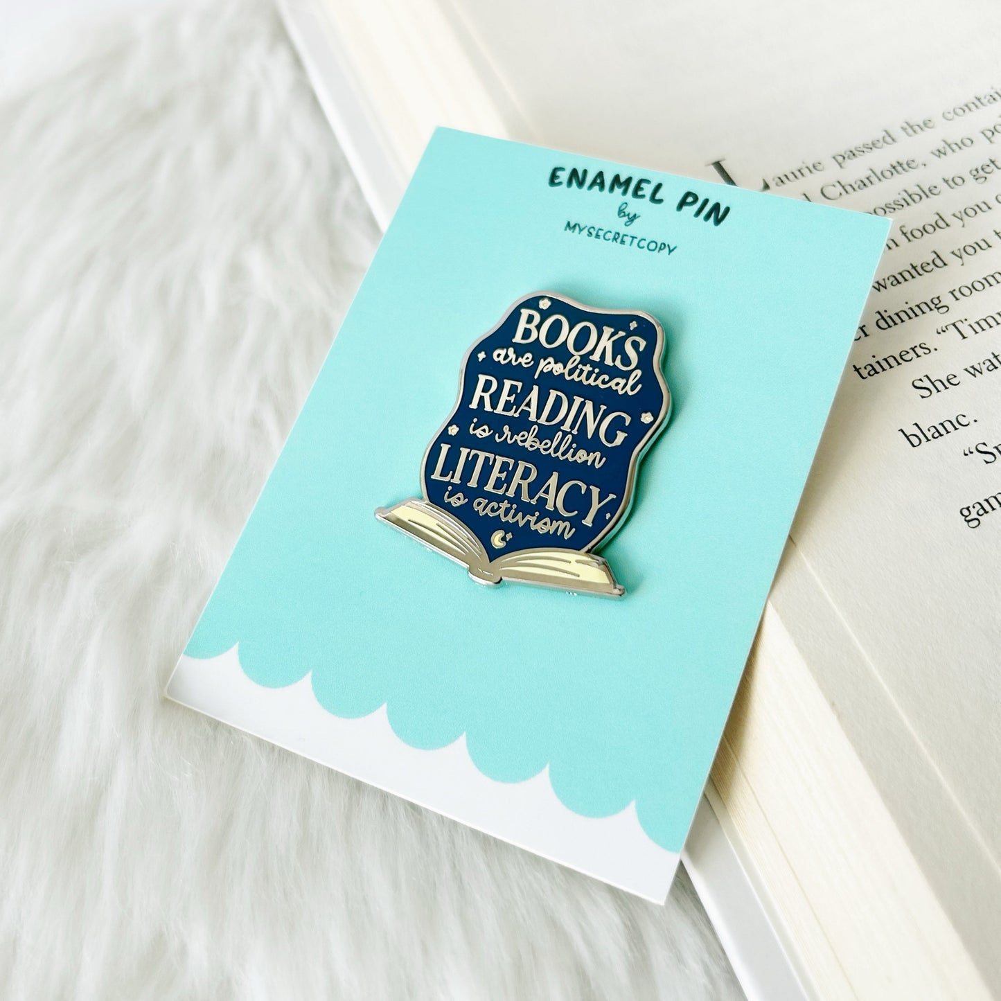 Enamel pin with book design on a matching card, placed on a surface with text.