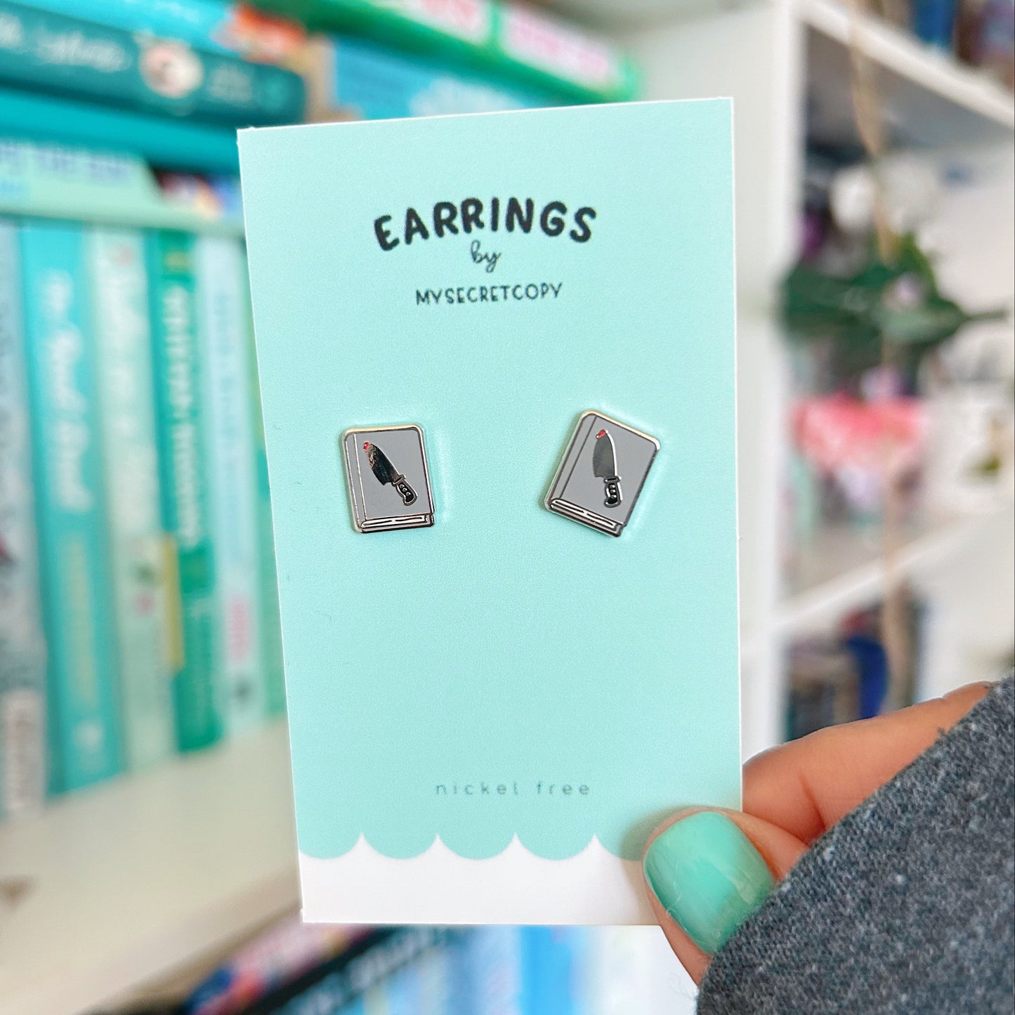 knife earrings on a card held in front of a blurred bookshelf