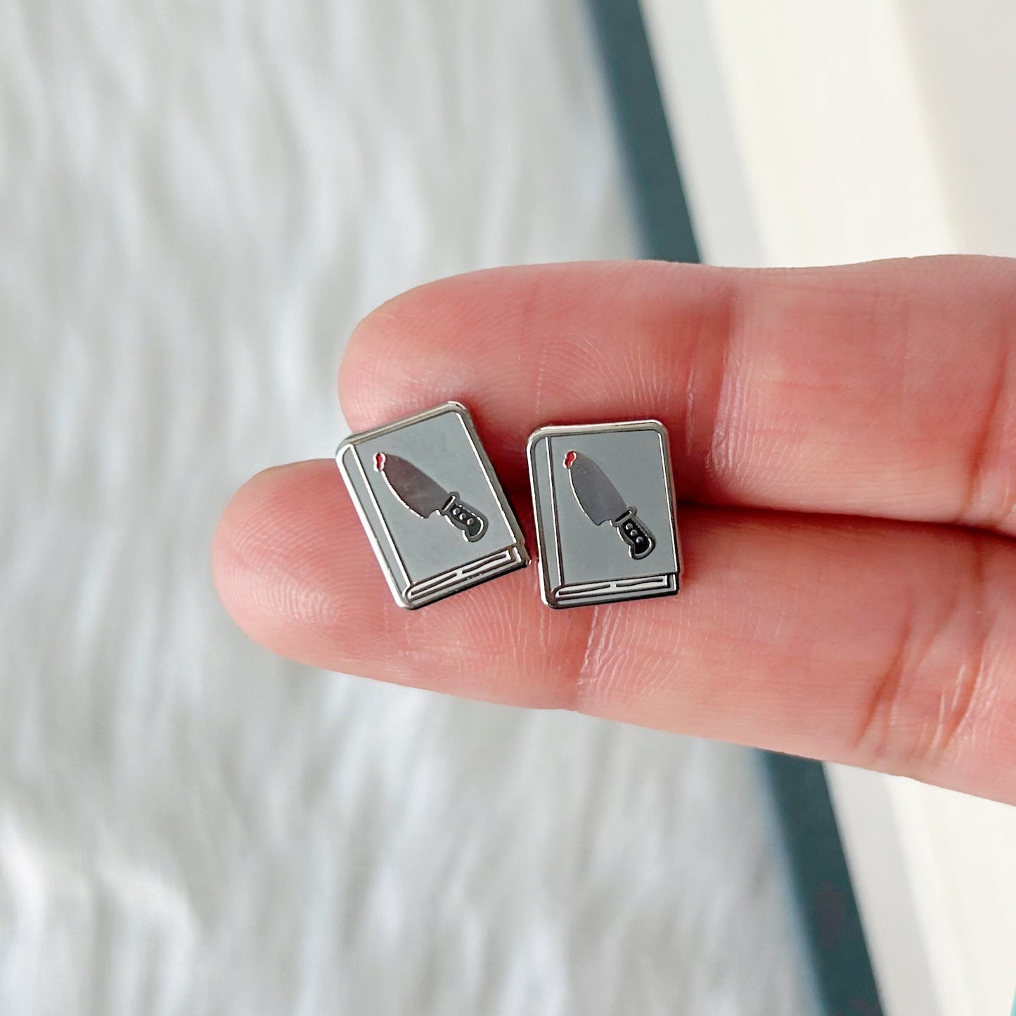 Person holding two square earrings with feather designs against a book background
