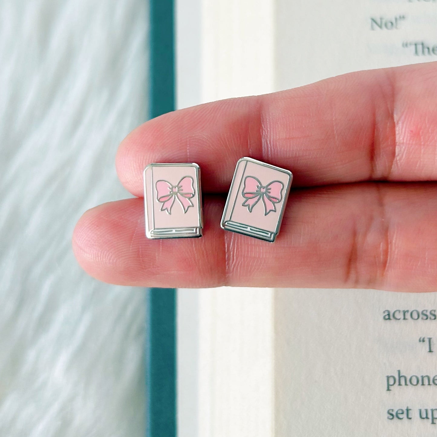 Hand holding two small square earrings with bow designs against a book background