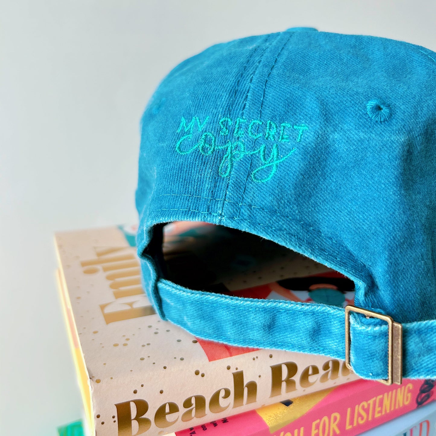 Blue cap with 'My Secret Copy' embroidery on a stack of books.