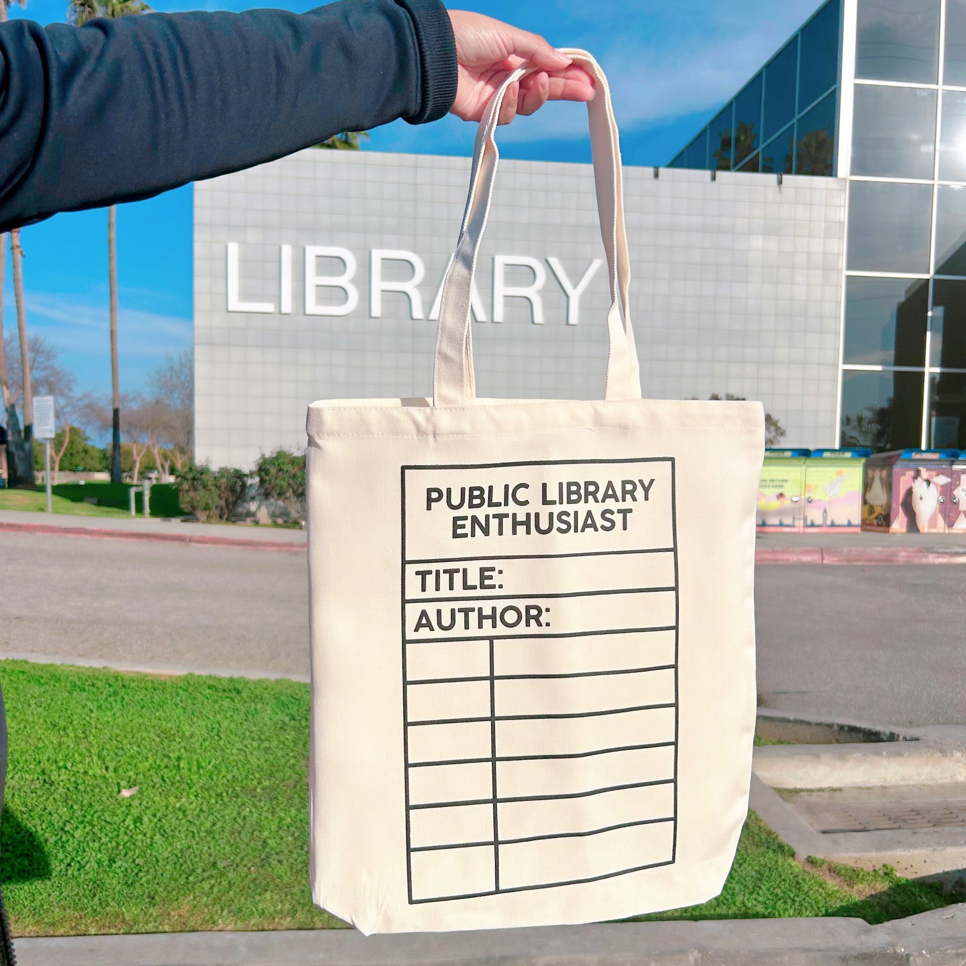 Tote bag with 'PUBLIC LIBRARY ENTHUSIAST' design held by a person outdoors.