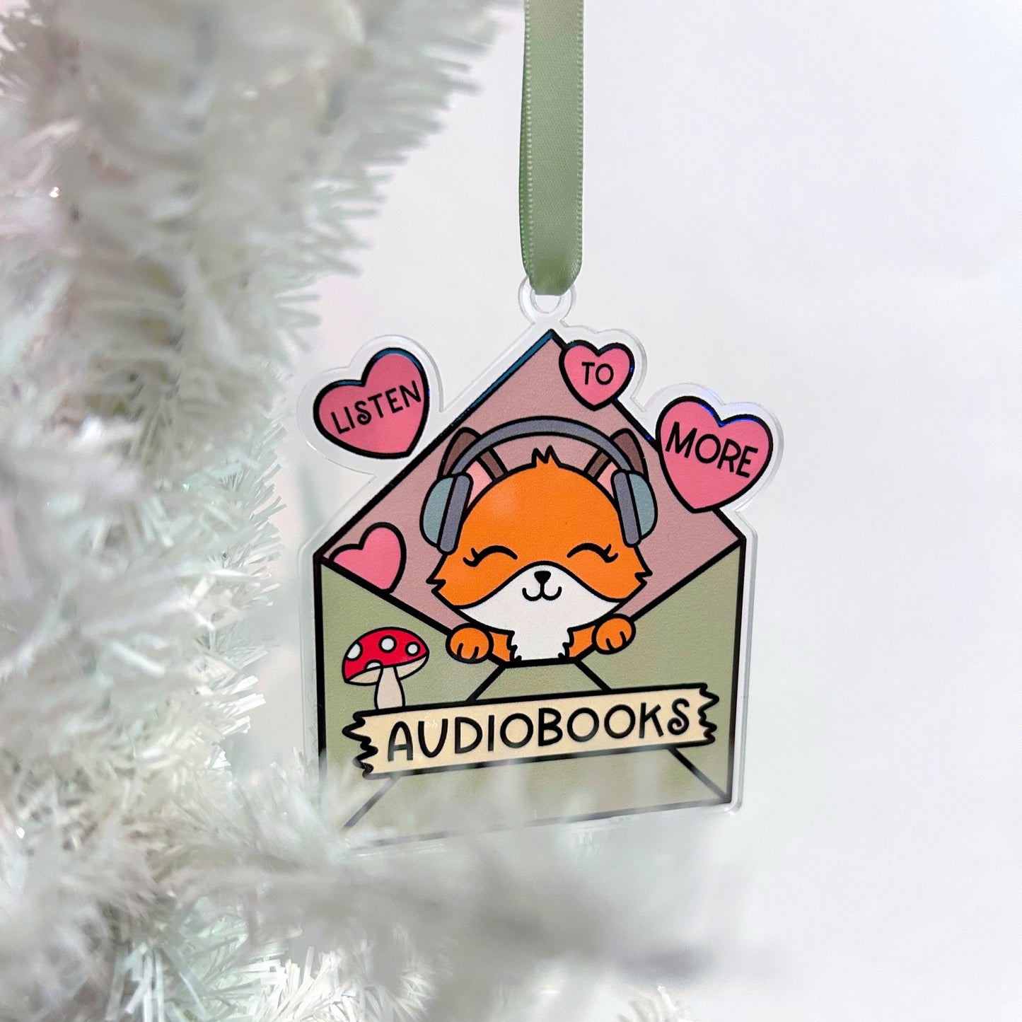 Decorative tag with a cartoon fox wearing headphones, surrounded by hearts and text 'Listen to More Audiobooks', hanging on a white Christmas tree.