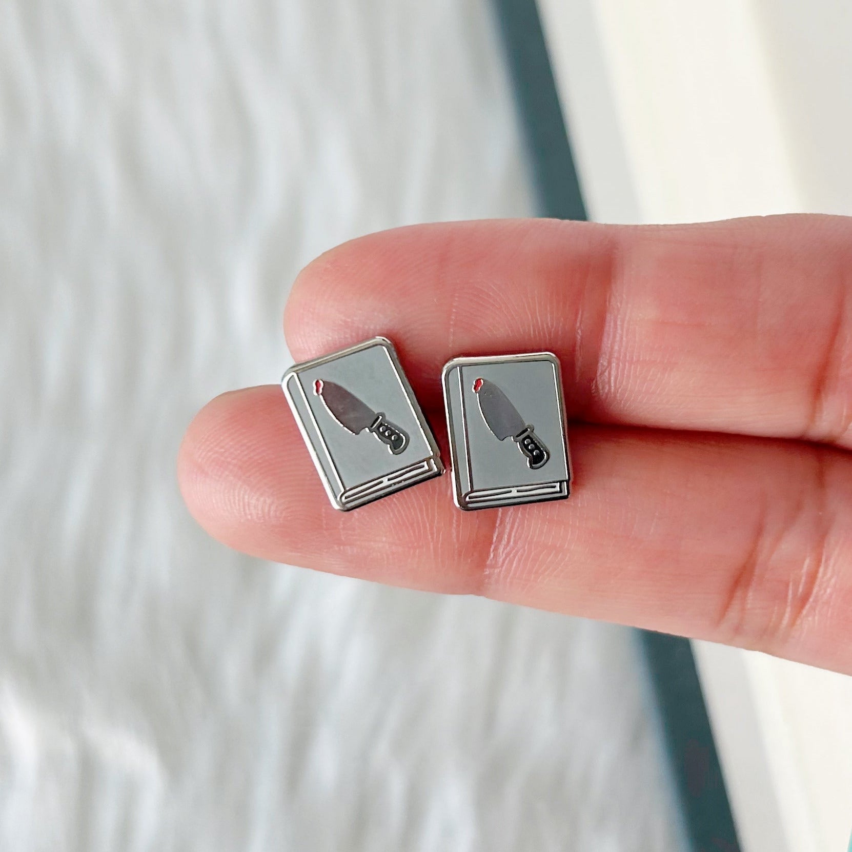 Person holding two square earrings with feather designs against a book background