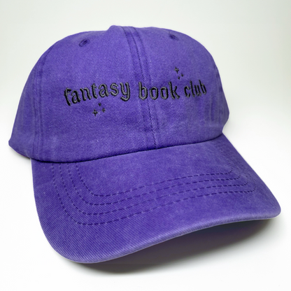 A purple cotton baseball cap with 'fantasy book club' embroidered in black gothic-style lettering on the front, set against a plain white background.
