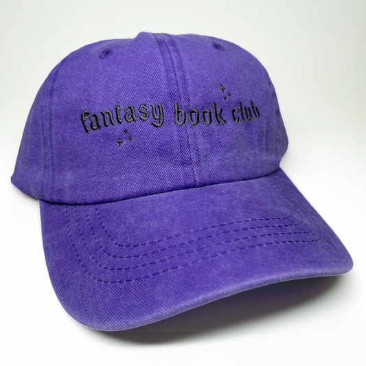 A purple cotton baseball cap with 'fantasy book club' embroidered in black gothic-style lettering on the front, set against a plain white background.