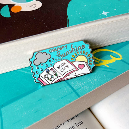 grumpy sunshine enamel pin on an open book with a character illustration above