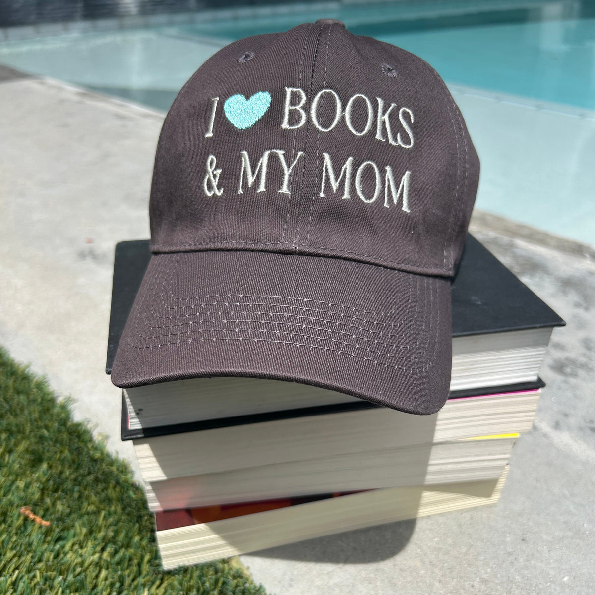 A dark brown baseball cap with 'I [heart] BOOKS & MY MOM' embroidered on the front, resting on a stack of books outdoors next to a swimming pool.