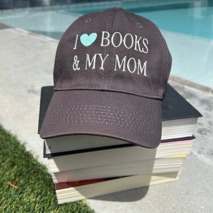 A dark brown baseball cap with 'I [heart] BOOKS & MY MOM' embroidered on the front, resting on a stack of books outdoors next to a swimming pool.
