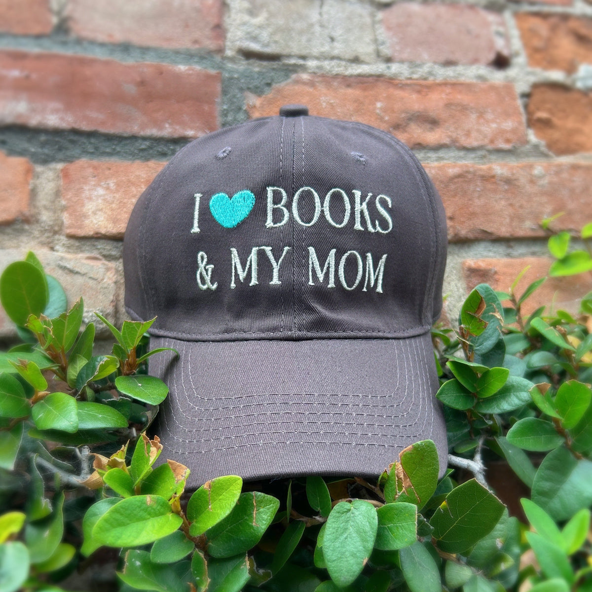 A baseball cap with 'I [heart] BOOKS & MY MOM' embroidered on the front, displayed outdoors among green leaves against a red brick wall.