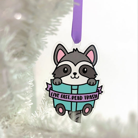 Decorative ornament with a raccoon design with a banner that reads "live fast, read trash" on a white Christmas tree.