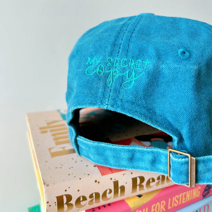 Blue cap with 'My Secret Copy' embroidery on a stack of books.