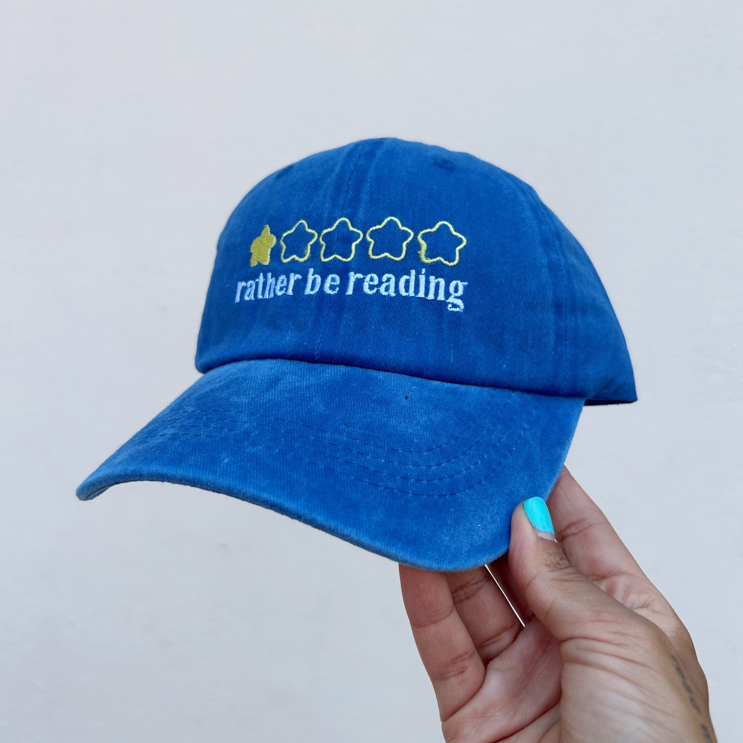 A blue cotton baseball cap held by a hand, featuring white embroidered text reading 'rather be reading' and a row of stars with one filled star in yellow.