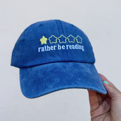 A blue washed baseball cap held by a hand against a plain light gray background. The front of the cap features white embroidered text reading 'rather be reading' below a row of five stars, with the first star filled in with yellow.