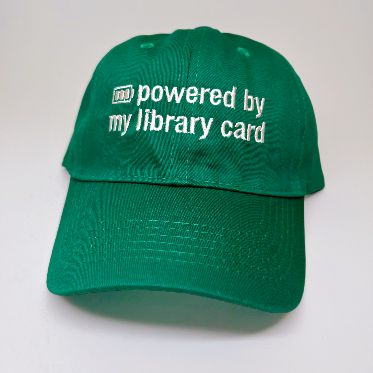 A green baseball cap with white embroidered text reading 'powered by my library card' on the front, photographed against a plain light background.