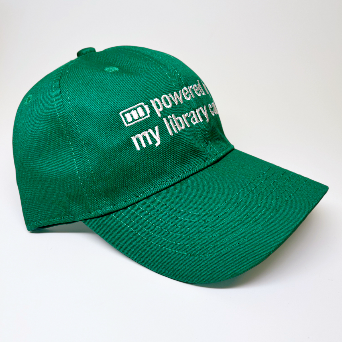 A green baseball cap with white embroidered text reading 'my library card' next to a small icon, photographed on a clean white background.