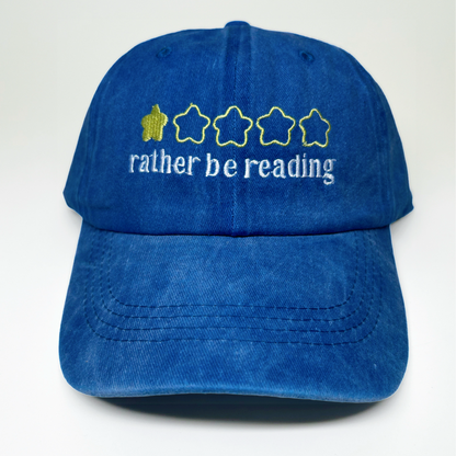 A blue cotton baseball cap with embroidered text reading 'rather be reading' and a yellow star graphic on the front, shown against a plain white background.