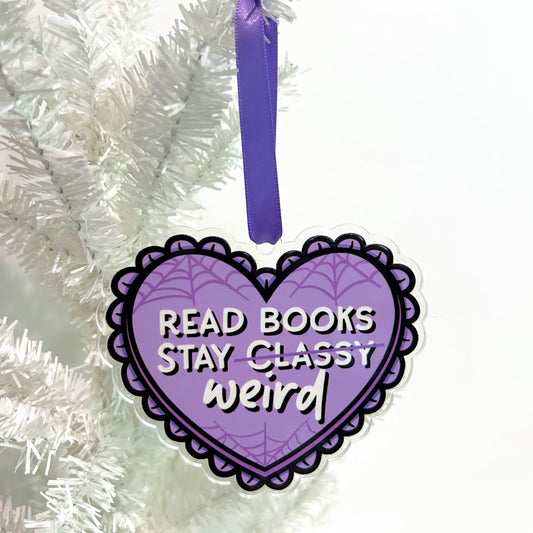 Purple heart-shaped ornament with text 'READ BOOKS STAY weird' hanging on a white Christmas tree.