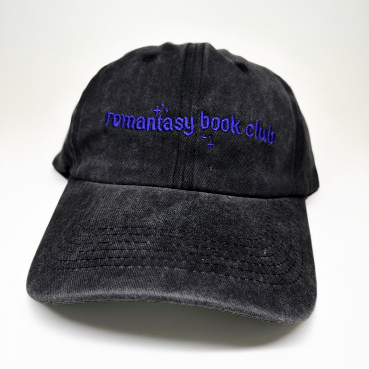 A black washed cotton baseball cap with purple embroidered text reading 'romantasy book club' on the front, shown against a plain white background.