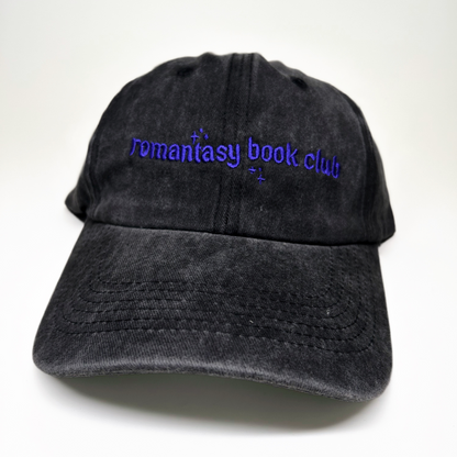 A black washed cotton baseball cap with purple embroidered text reading 'romantasy book club' on the front, shown against a plain white background.
