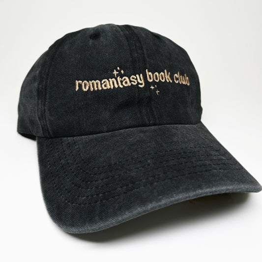A black washed cotton baseball cap with 'romantasy book club' embroidered in gold across the front, set against a plain background.