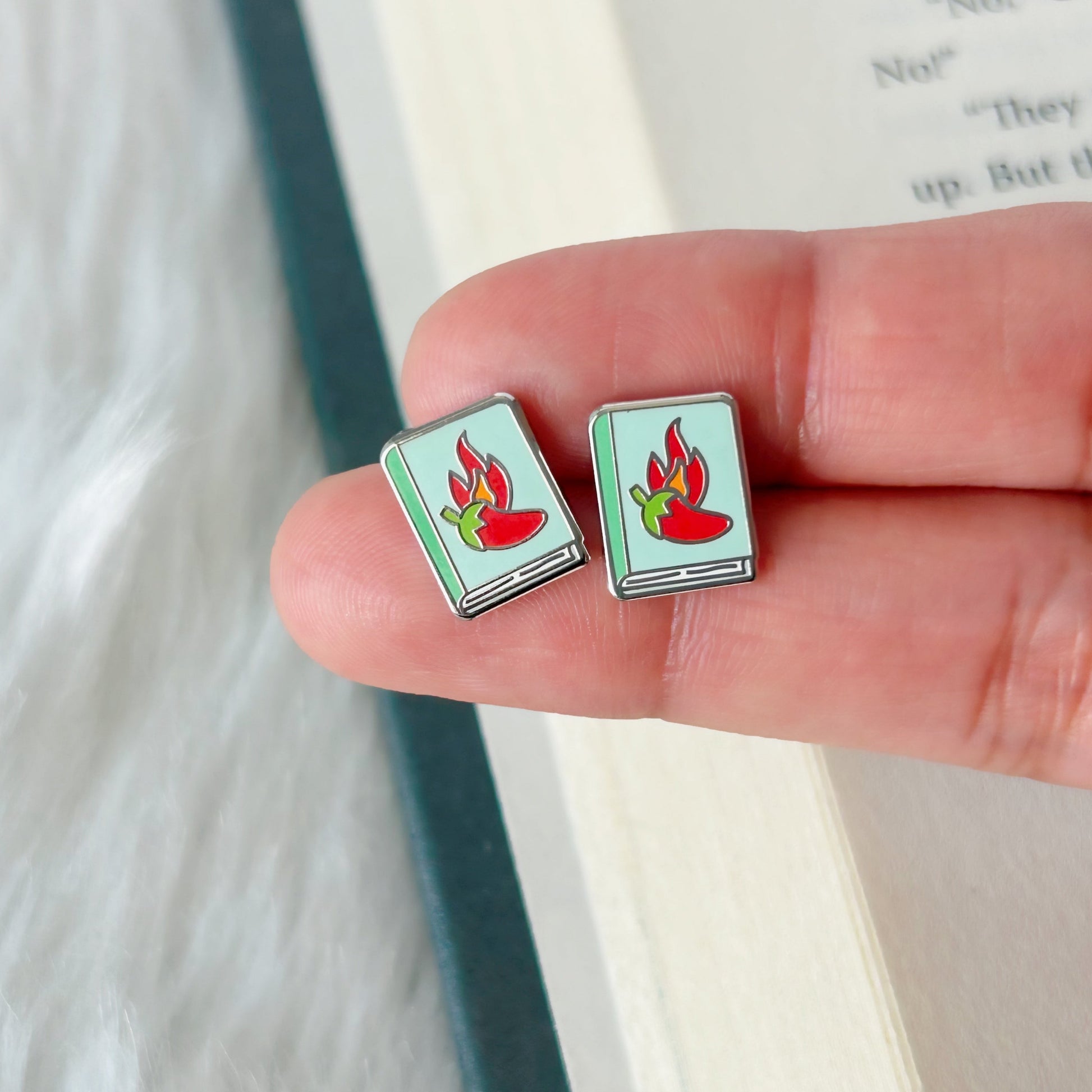 Square earrings with a red and green design held between fingers against a blurred book background
